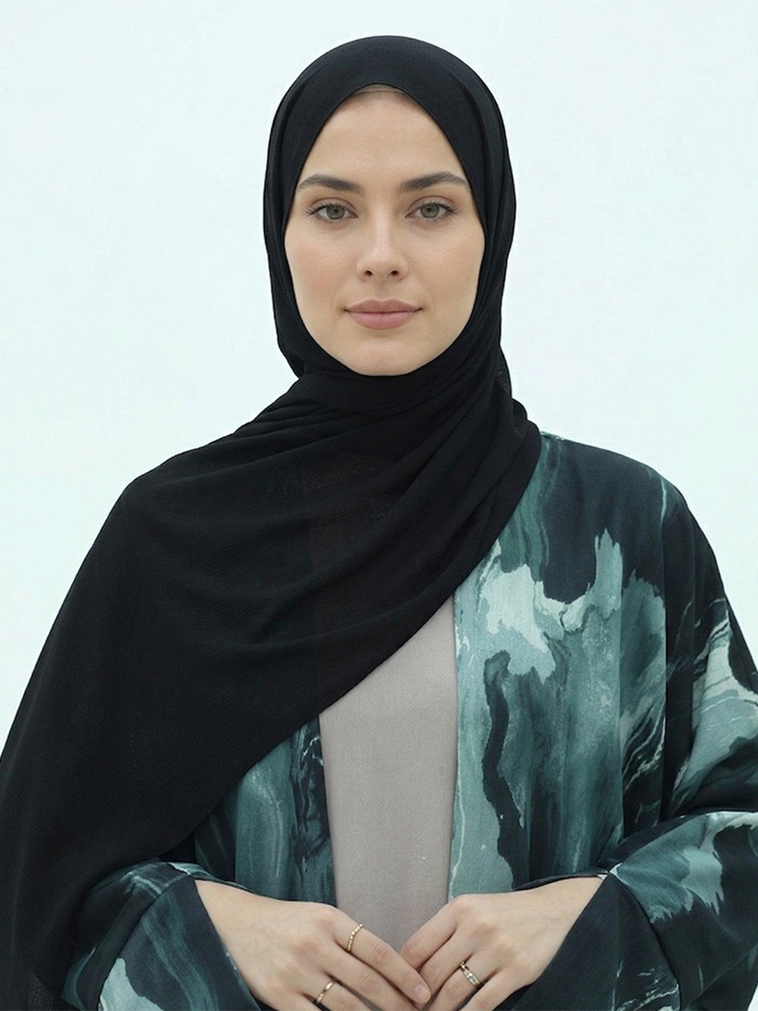 A woman wearing a black hijab and a long-sleeved abaya with a green and black abstract watercolor pattern over a light gray inner top, set against a plain light gray background.