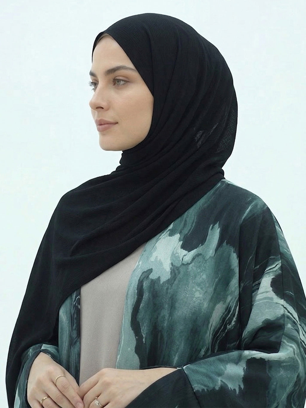 A woman wearing a black hijab and a long-sleeved abstract print abaya in shades of green, gray, and black, posed against a light off-white background.
