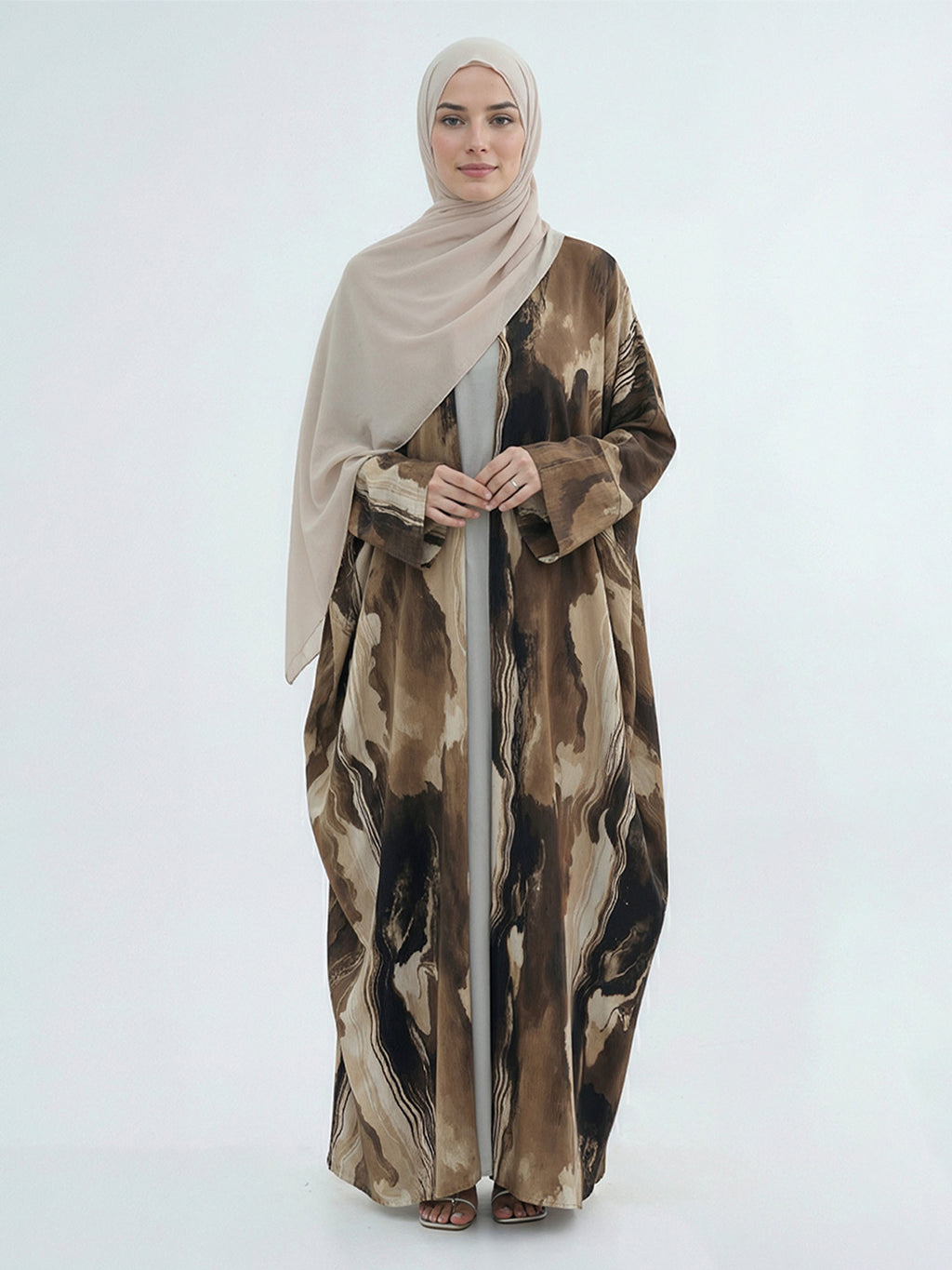 A woman wearing a beige hijab and a long, flowing abaya with an abstract brown, beige, and black watercolor-style print, standing against a plain light gray studio background.