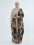 A woman wearing a beige hijab and a long, flowing abaya with an abstract brown, beige, and black watercolor-style print, standing against a plain light gray studio background.