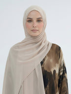 A woman wearing a light beige chiffon hijab and a brown and black patterned blouse, photographed against a clean light gray studio background.