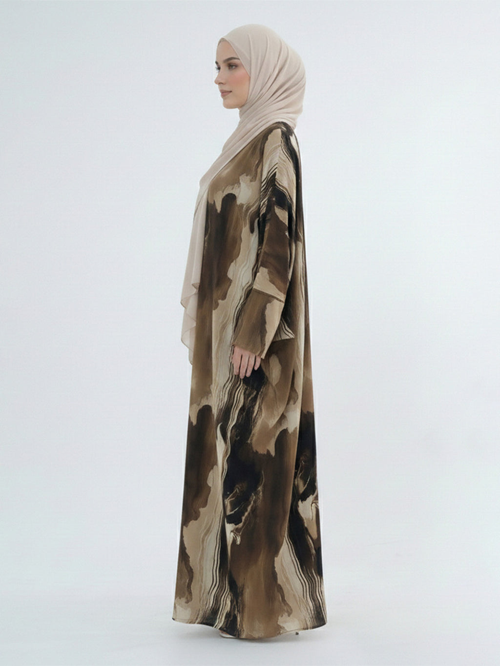 A woman in profile wearing a long beige hijab and a flowing abaya with an abstract brown and beige watercolor-style print, photographed against a plain light gray studio background.