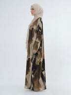 A woman in profile wearing a long beige hijab and a flowing abaya with an abstract brown and beige watercolor-style print, photographed against a plain light gray studio background.