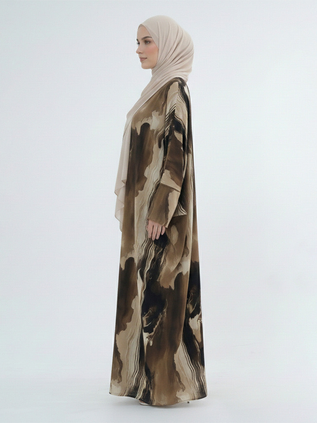 A woman in profile wearing a long beige hijab and a flowing abaya with an abstract brown and beige watercolor-style print, photographed against a plain light gray studio background.