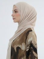 A woman in profile wearing a light beige hijab and a brown abstract-printed blouse against a plain light gray background.
