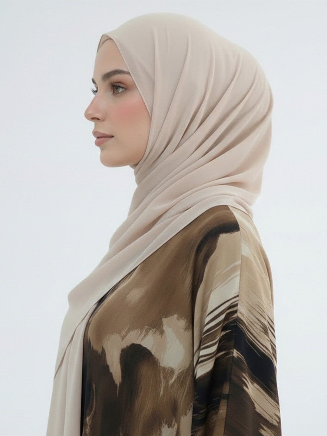 A woman in profile wearing a light beige hijab and a brown abstract-printed blouse against a plain light gray background.