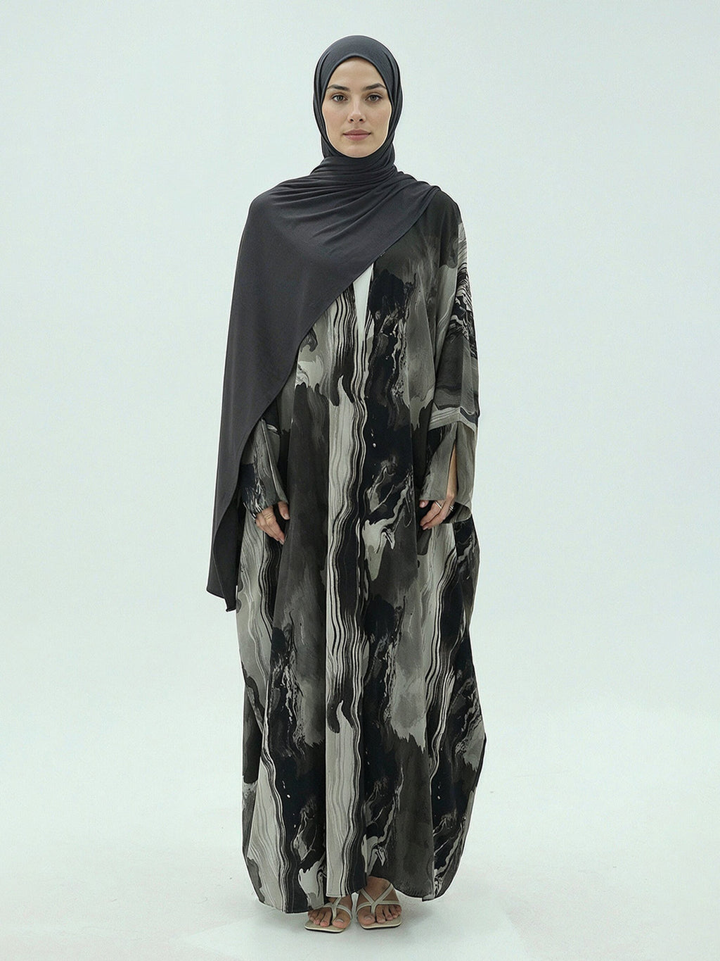 A woman modeling a long-sleeved, floor-length abaya with a black, gray, and white abstract watercolor pattern, paired with a solid black hijab, against a light gray studio background.