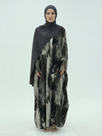 A woman modeling a long-sleeved, floor-length abaya with a black, gray, and white abstract watercolor pattern, paired with a solid black hijab, against a light gray studio background.