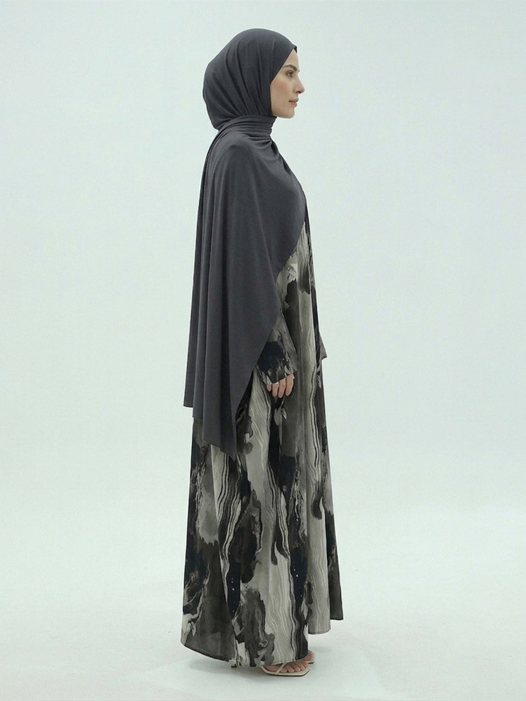A woman in profile wearing a dark gray hijab and a long, flowing abaya with a black, gray, and white abstract watercolor print, photographed against a light gray studio background.
