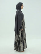 A woman in profile wearing a dark gray hijab and a long, flowing abaya with a black, gray, and white abstract watercolor print, photographed against a light gray studio background.
