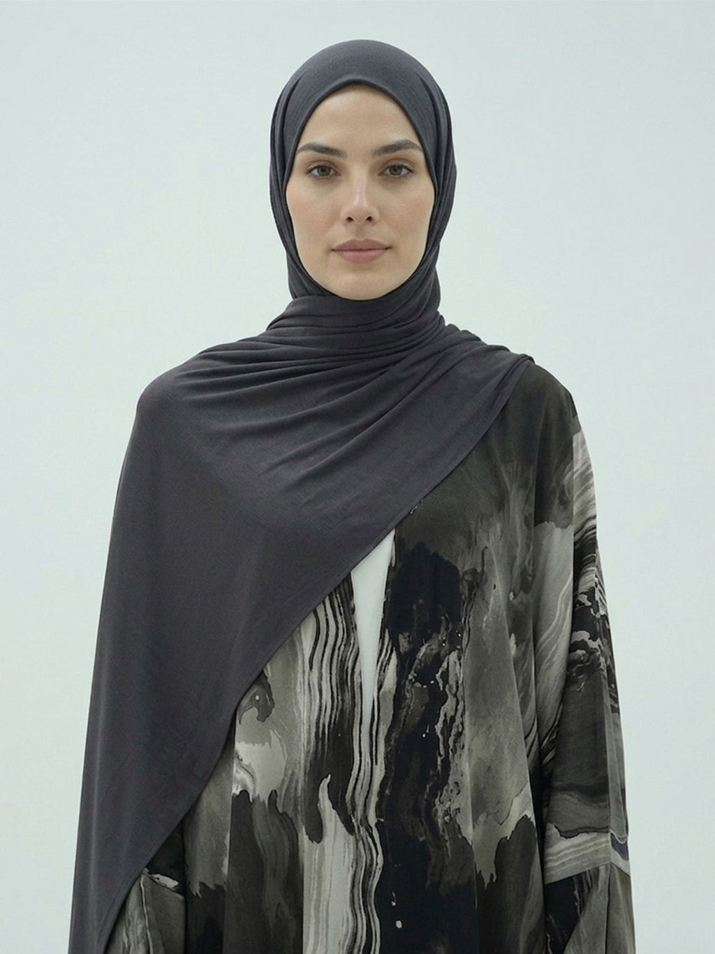 A woman wearing a dark charcoal gray hijab and a black, gray, and white abstract print abaya against a light gray studio background.