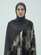A woman wearing a dark charcoal gray hijab and a black, gray, and white abstract print abaya against a light gray studio background.