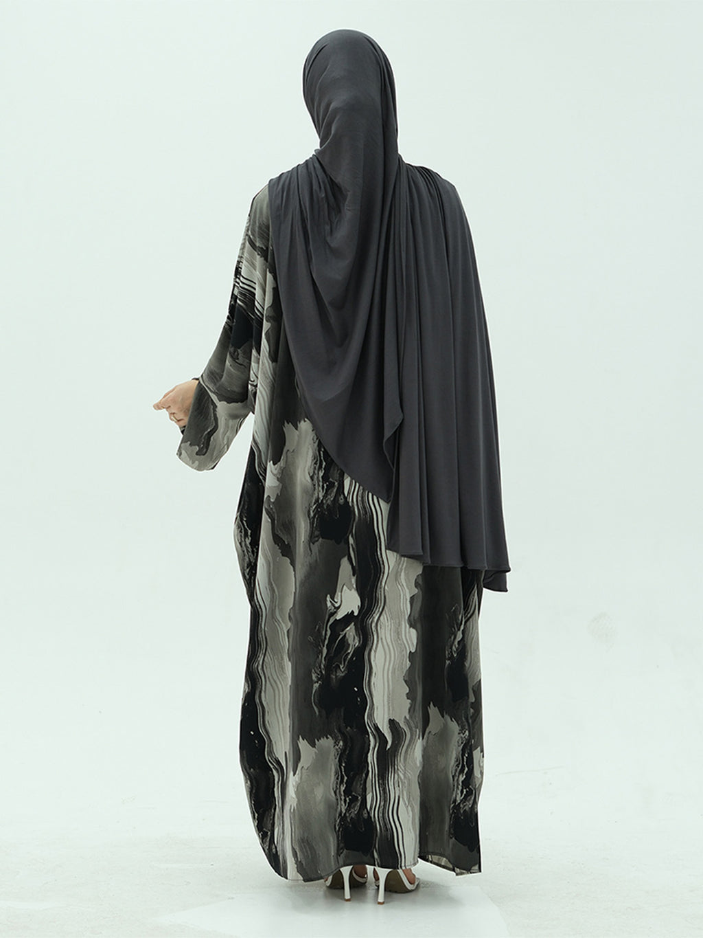 A woman shown from the back wearing a long black hijab and a long-sleeved abaya with a black, gray, and white abstract marbled pattern, set against a plain light gray studio background.