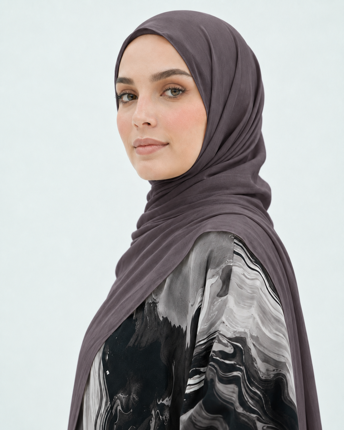A woman wearing a draped hijab in a muted dark plum-gray color, paired with a black, gray, and white marbled patterned top, shown against a light gray studio background.