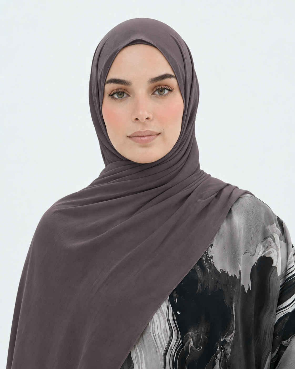 A woman wearing a draped taupe-brown hijab and a black and gray patterned top, photographed against a clean light gray studio background.