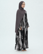 A woman modeling a modest long-sleeve maxi dress with a black, gray, and white abstract watercolor print, paired with a solid dark brown hijab, against a light gray studio background.