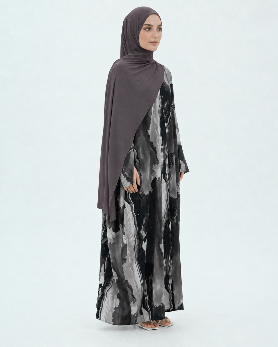 A woman modeling a modest long-sleeve maxi dress with a black, gray, and white abstract watercolor print, paired with a solid dark brown hijab, against a light gray studio background.
