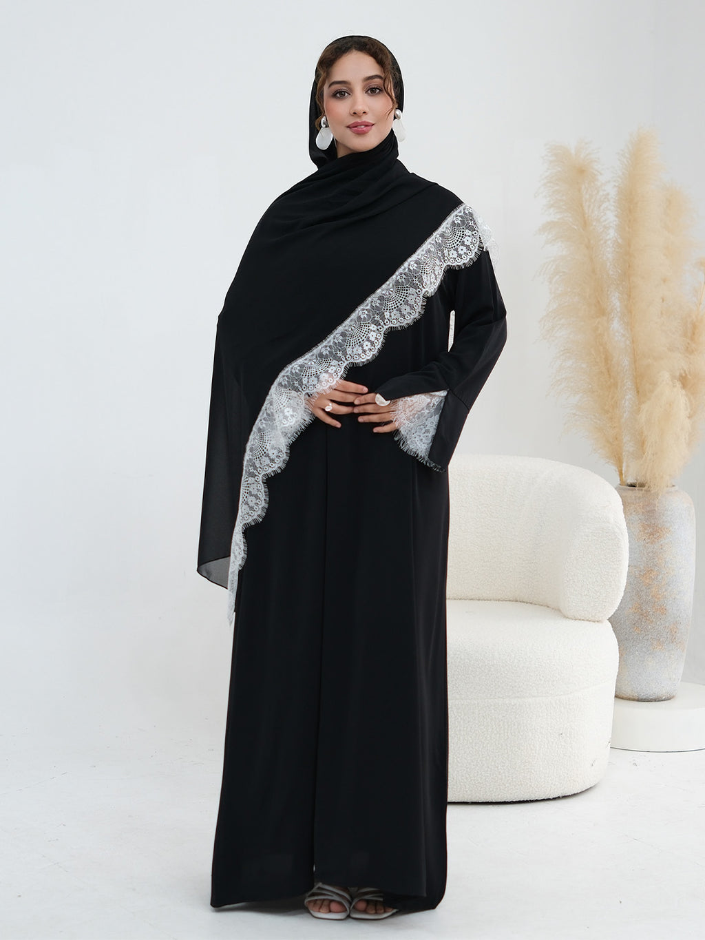 A woman modeling a black abaya with a wide silver-gray lace-trimmed sash draped diagonally across the chest, standing in a bright studio with a white curved chair and pampas grass in the background.