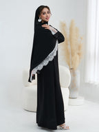 A woman modeling a black long abaya with silver lace trim, paired with a matching black hijab, standing in a bright, modern room with a white chair and pampas grass.