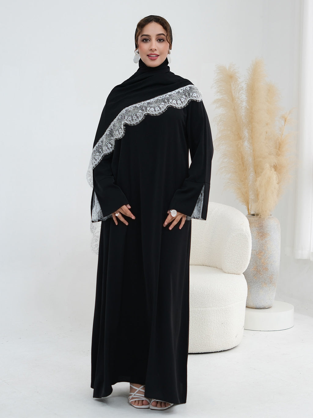 A woman modeling a long black abaya dress with a wide silver-gray lace draped shoulder detail, standing in a bright studio with a white curved chair and pampas grass decor in the background.