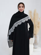 A woman modeling a black abaya dress with a wide, decorative silver lace-trimmed shawl draped over her shoulders, set against a clean white studio background with pampas grass decor.