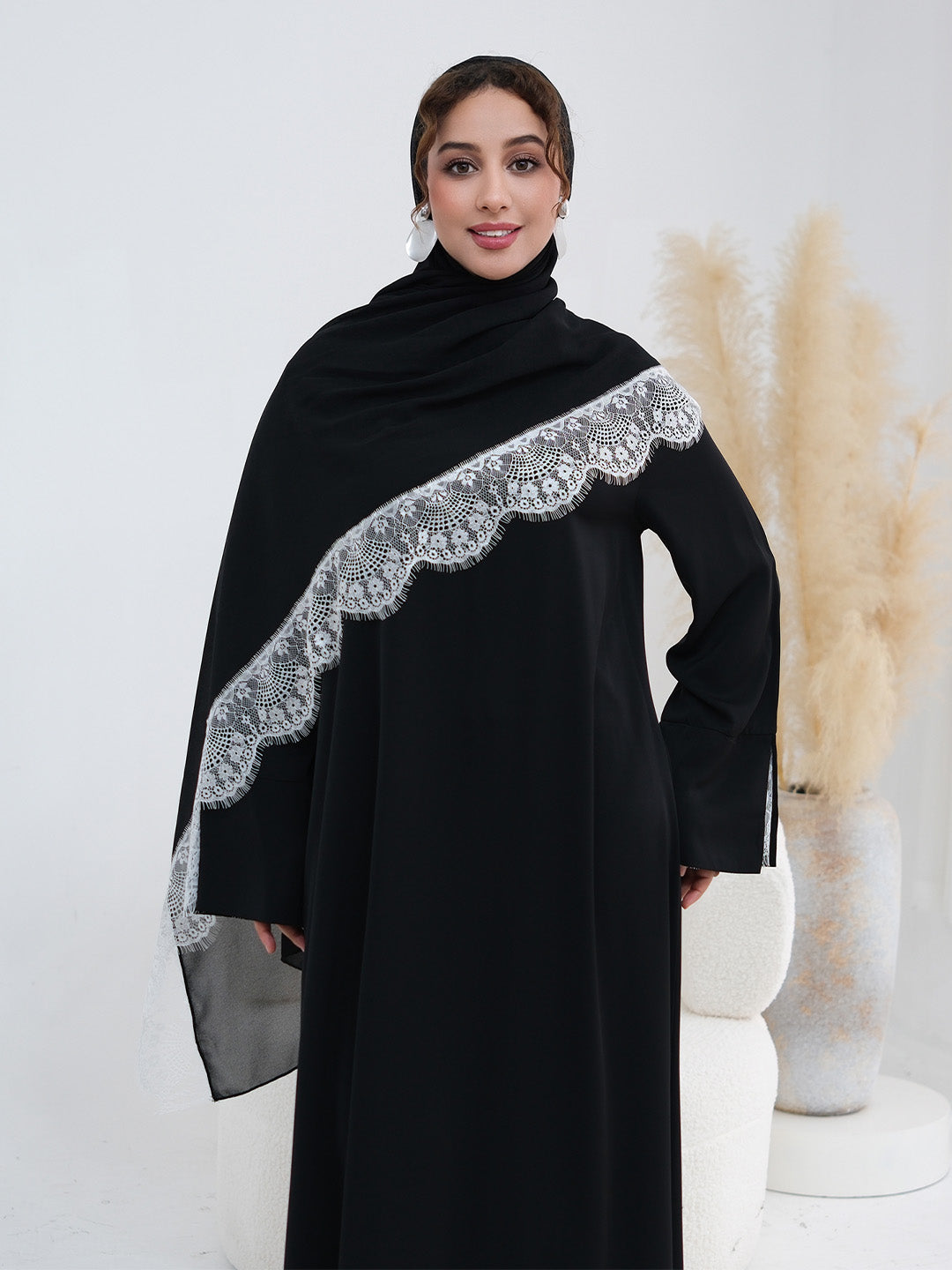 A woman modeling a black abaya dress with a wide, decorative silver lace-trimmed shawl draped over her shoulders, set against a clean white studio background with pampas grass decor.