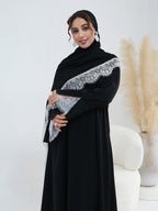 A woman wearing a black abaya with a wide black shawl featuring silver-gray lace trim, posed in front of a white studio background with a decorative vase of pampas grass.