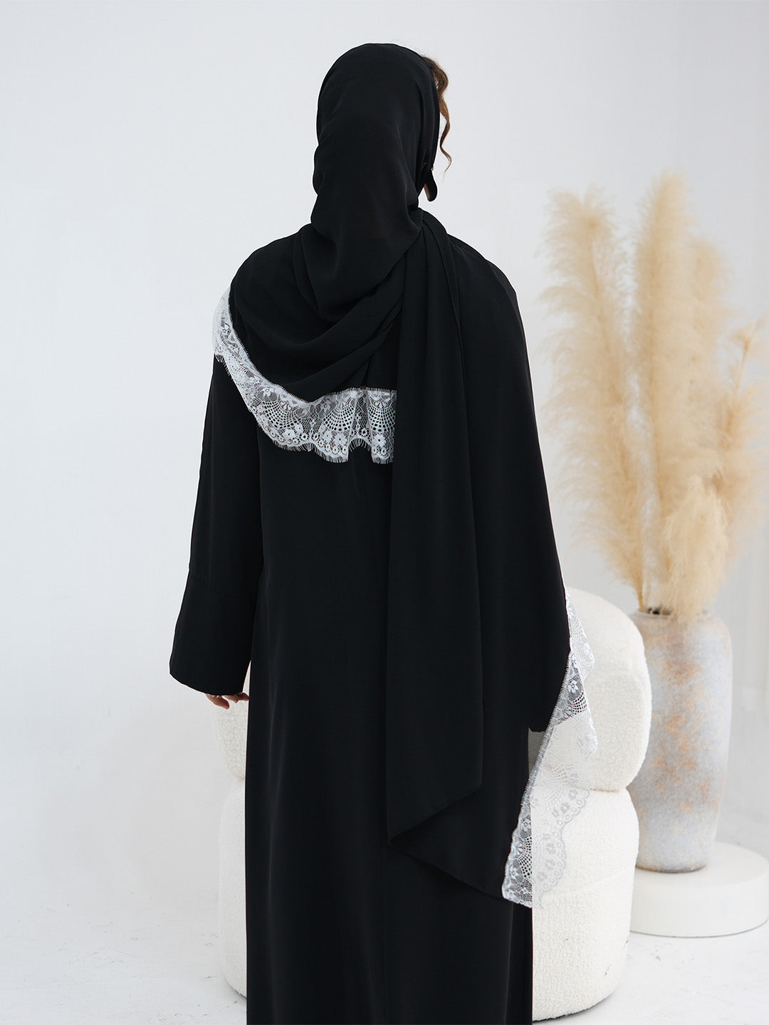 A woman shown from the back wearing a black abaya with a wide silver-gray lace trim detail across the shoulders and along the sleeve cuffs, standing in a bright studio with decorative pampas grass.