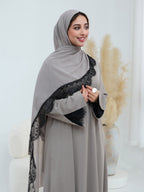 A woman wearing a light gray abaya and matching hijab with black lace trim, standing in a bright indoor setting with decorative pampas grass in the background.