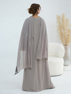 A woman shown from the back wearing a flowing, floor-length taupe-gray abaya with a matching sheer draped overlay, standing in a bright studio next to a white curved chair and a vase of pampas grass.