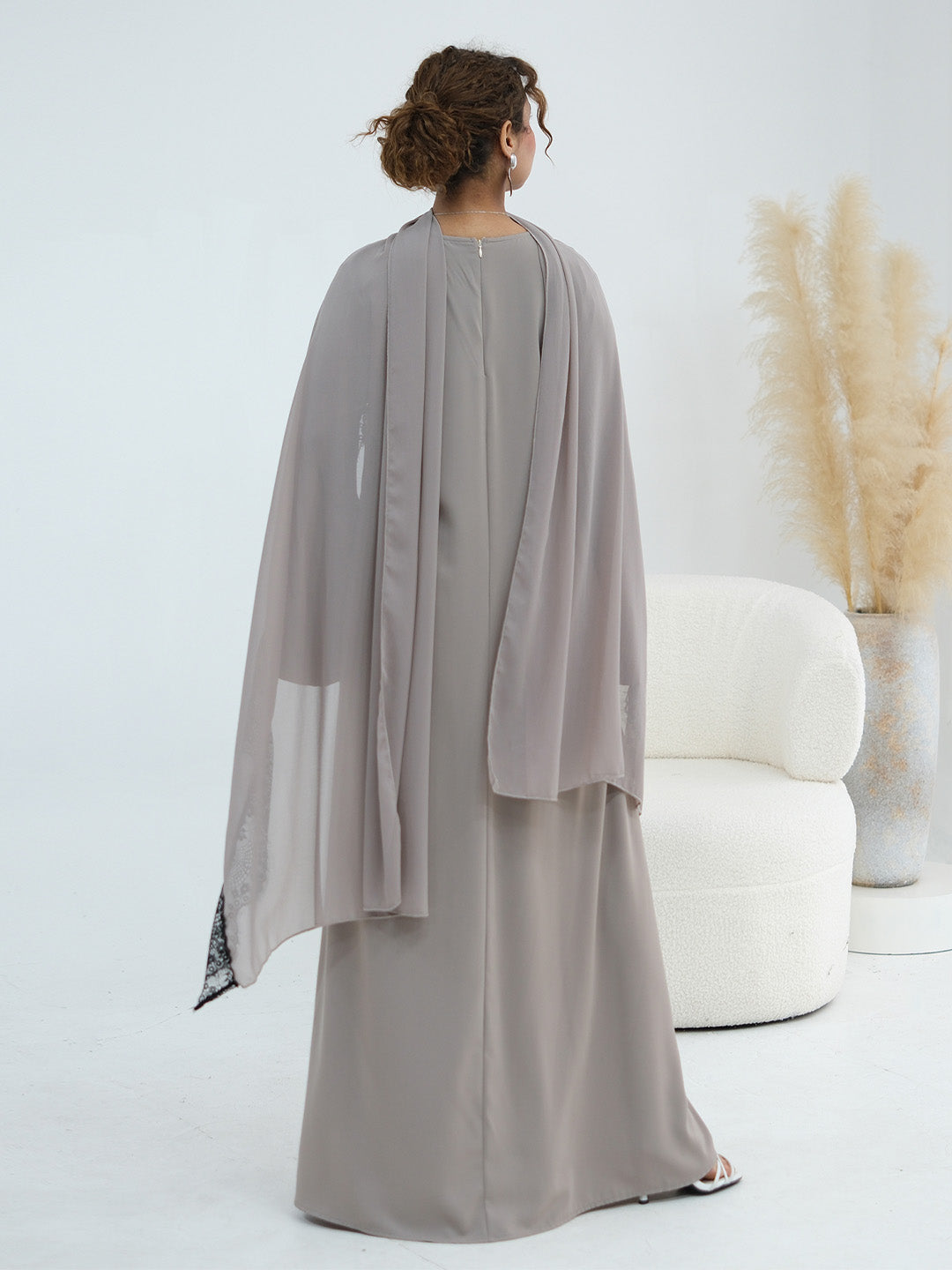 A woman shown from the back wearing a flowing, floor-length taupe-gray abaya with a matching sheer draped overlay, standing in a bright studio next to a white curved chair and a vase of pampas grass.