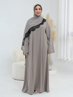 A woman wearing a long taupe-gray abaya with black lace trim along the shoulder, paired with a matching hijab, standing in a bright, modern room with a white chair and pampas grass.