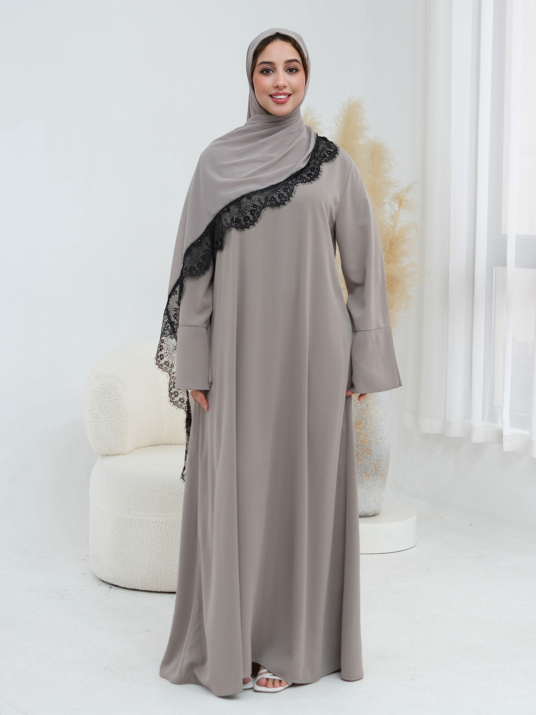 A woman wearing a long taupe-gray abaya with black lace trim along the shoulder, paired with a matching hijab, standing in a bright, modern room with a white chair and pampas grass.