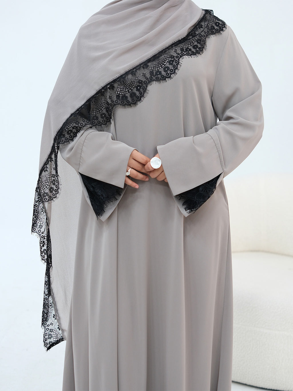 A woman wearing a long-sleeved light gray abaya with black lace trim along the shoulder and black lace cuffs, paired with a matching gray hijab, standing against a white background.