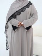 A woman wearing a long-sleeved light gray abaya with black lace trim along the shoulder and black lace cuffs, paired with a matching gray hijab, standing against a white background.