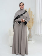 A woman wearing a floor-length taupe abaya dress with black lace trim along the shoulder and black fabric-covered cuffs, paired with a matching hijab, standing in a bright white interior with decorative pampas grass.