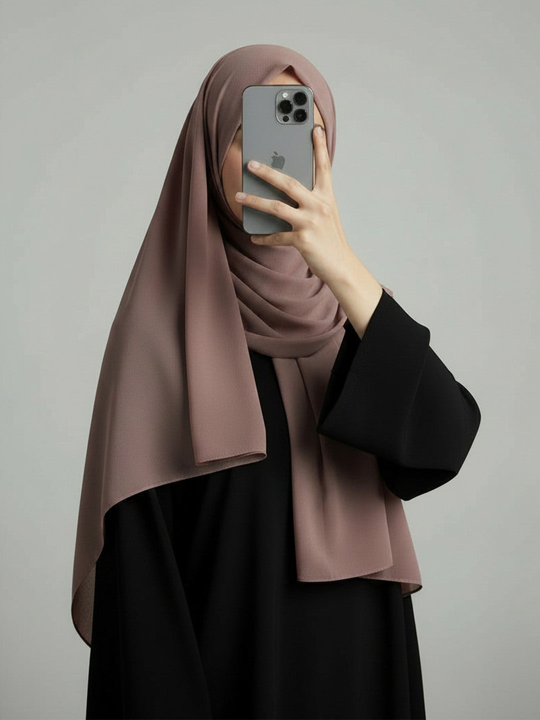 A woman wearing a draped dusty rose hijab and a black abaya takes a mirror selfie with a gray smartphone against a plain light gray background.