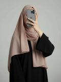 A woman wearing a dusty pink hijab and a black abaya takes a mirror selfie with her phone against a plain gray studio background.