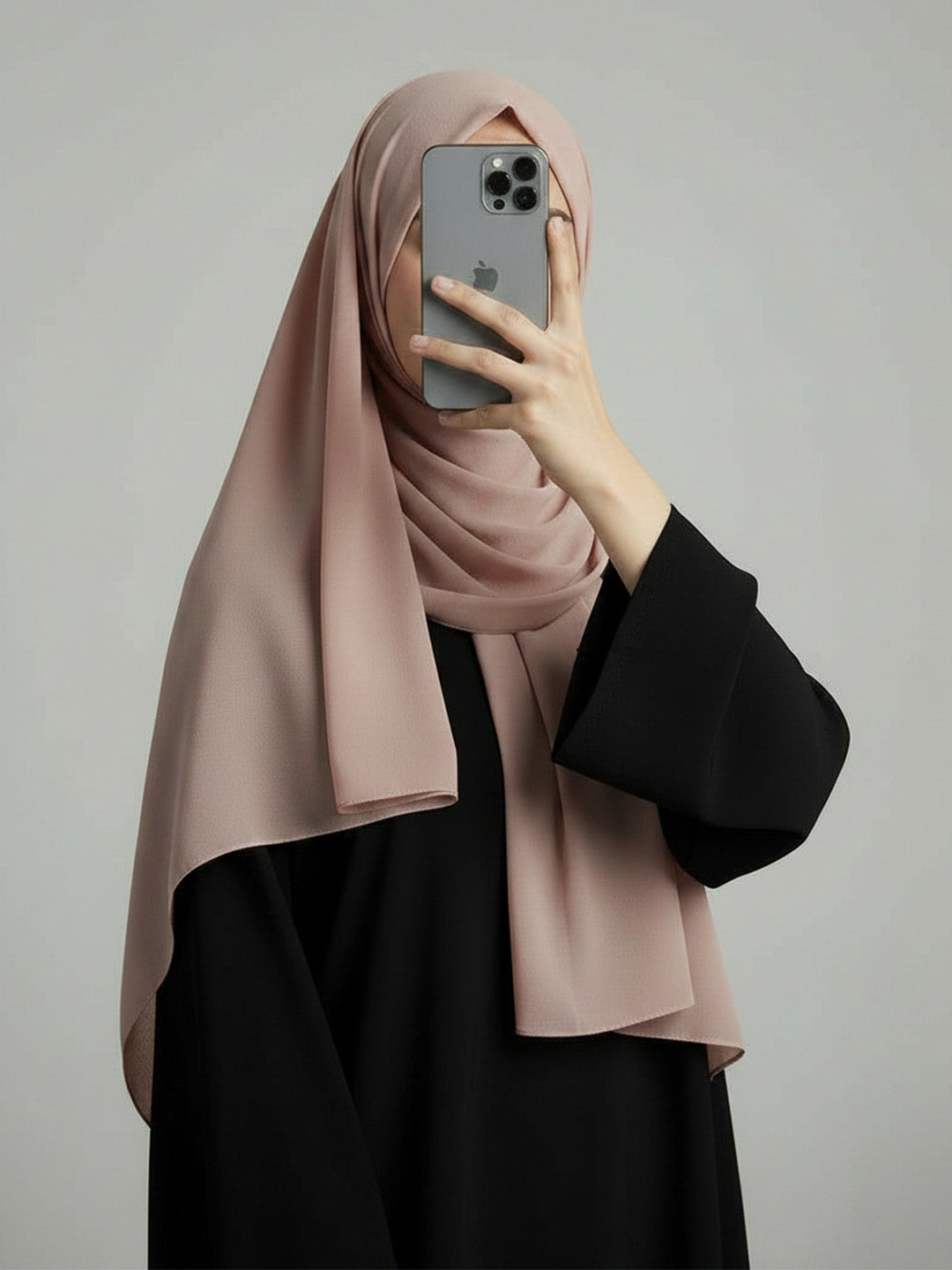 A woman wearing a dusty pink hijab and a black abaya takes a mirror selfie with her phone against a plain gray studio background.