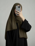 A woman wearing a dark olive green hijab and a black abaya takes a mirror selfie with her phone against a plain gray studio background.