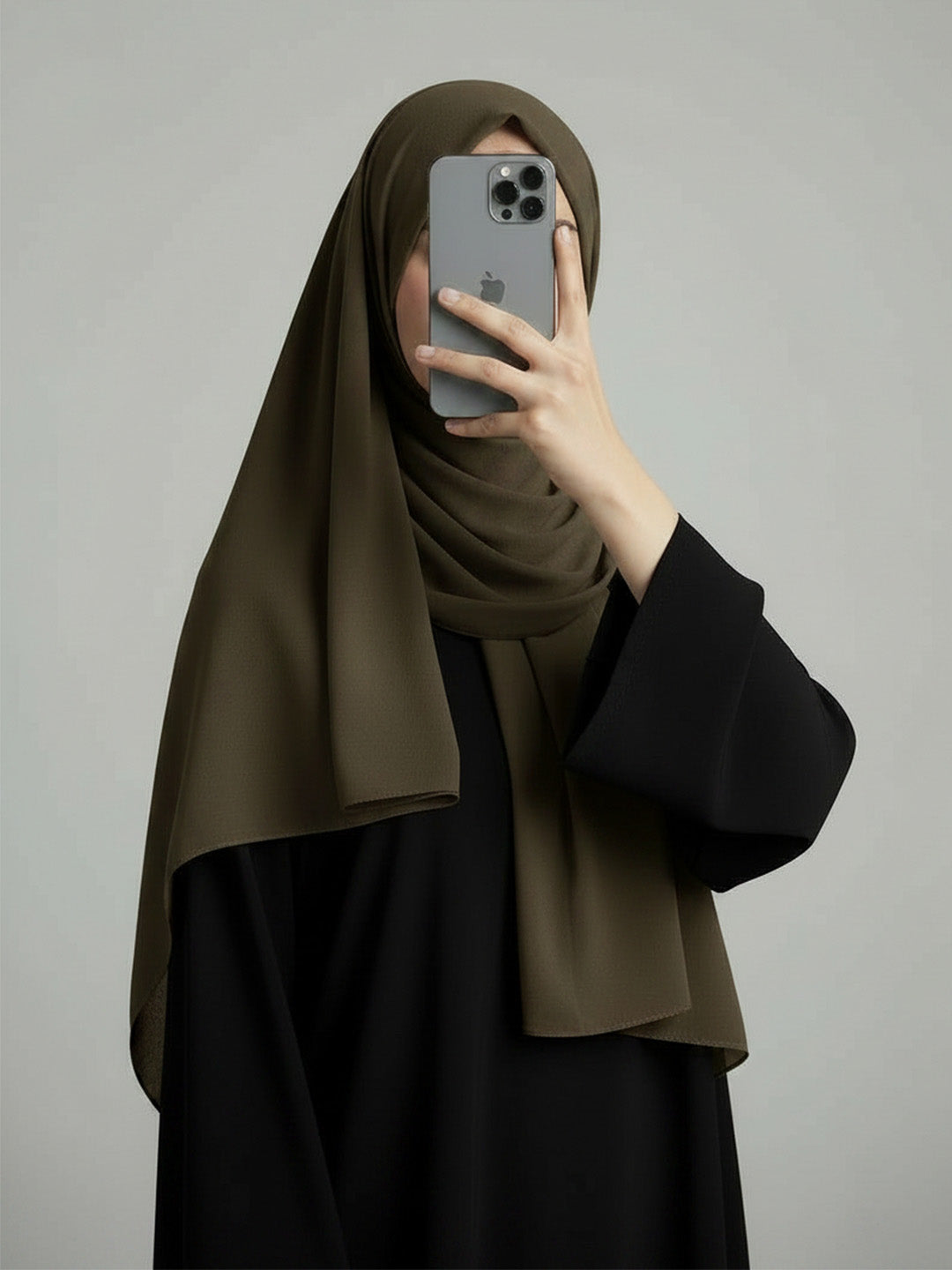 A woman wearing a dark olive green hijab and a black abaya takes a mirror selfie with her phone against a plain gray studio background.
