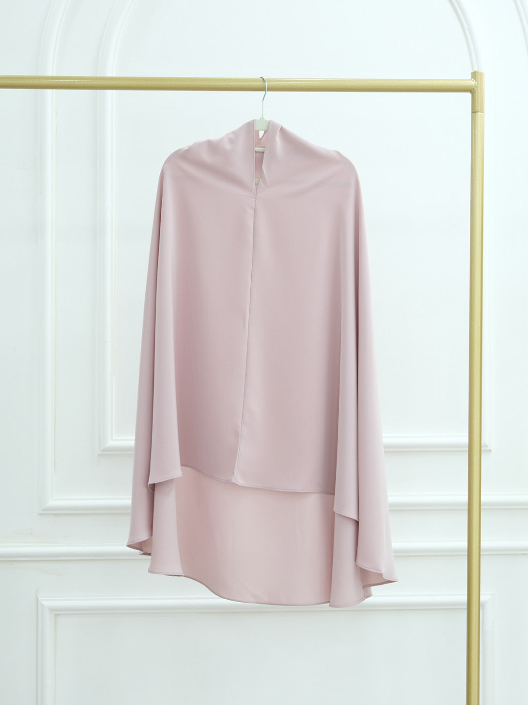 A dusty pink high-neck draped blouse with long flowing sleeves, hanging on a gold clothing rack against a white paneled wall.