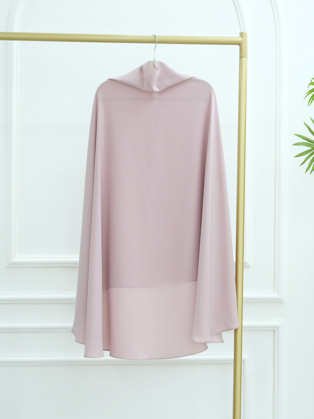 A light pink hooded cape-style top or dress displayed on a gold clothing rack against a white paneled wall.