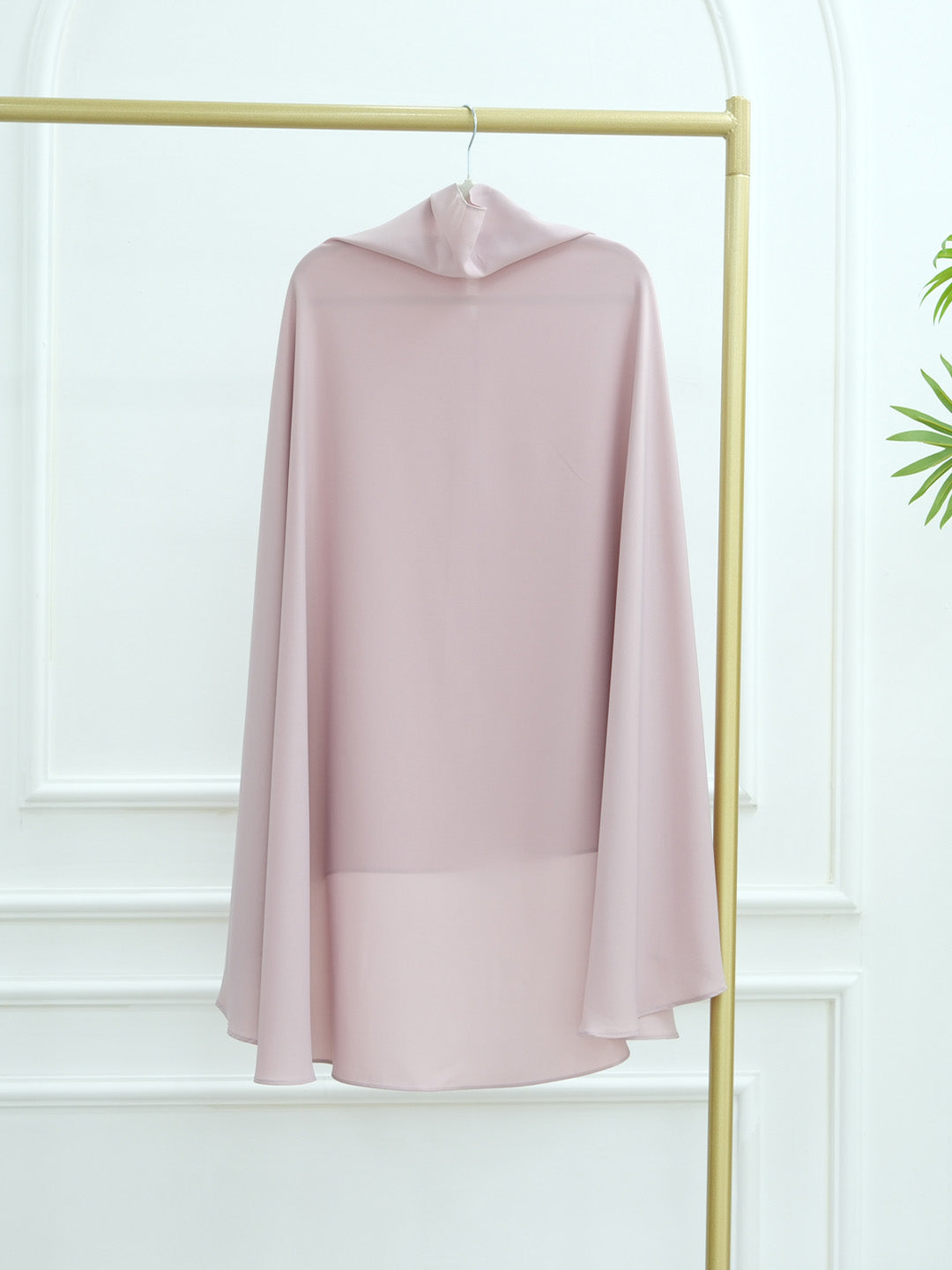 A light pink hooded cape-style top or dress displayed on a gold clothing rack against a white paneled wall.