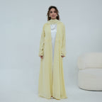 A woman stands in a studio wearing a long, flowing pale yellow abaya with a matching headscarf and a white inner dress visible underneath. A light upholstered chair is partially visible at the right edge.