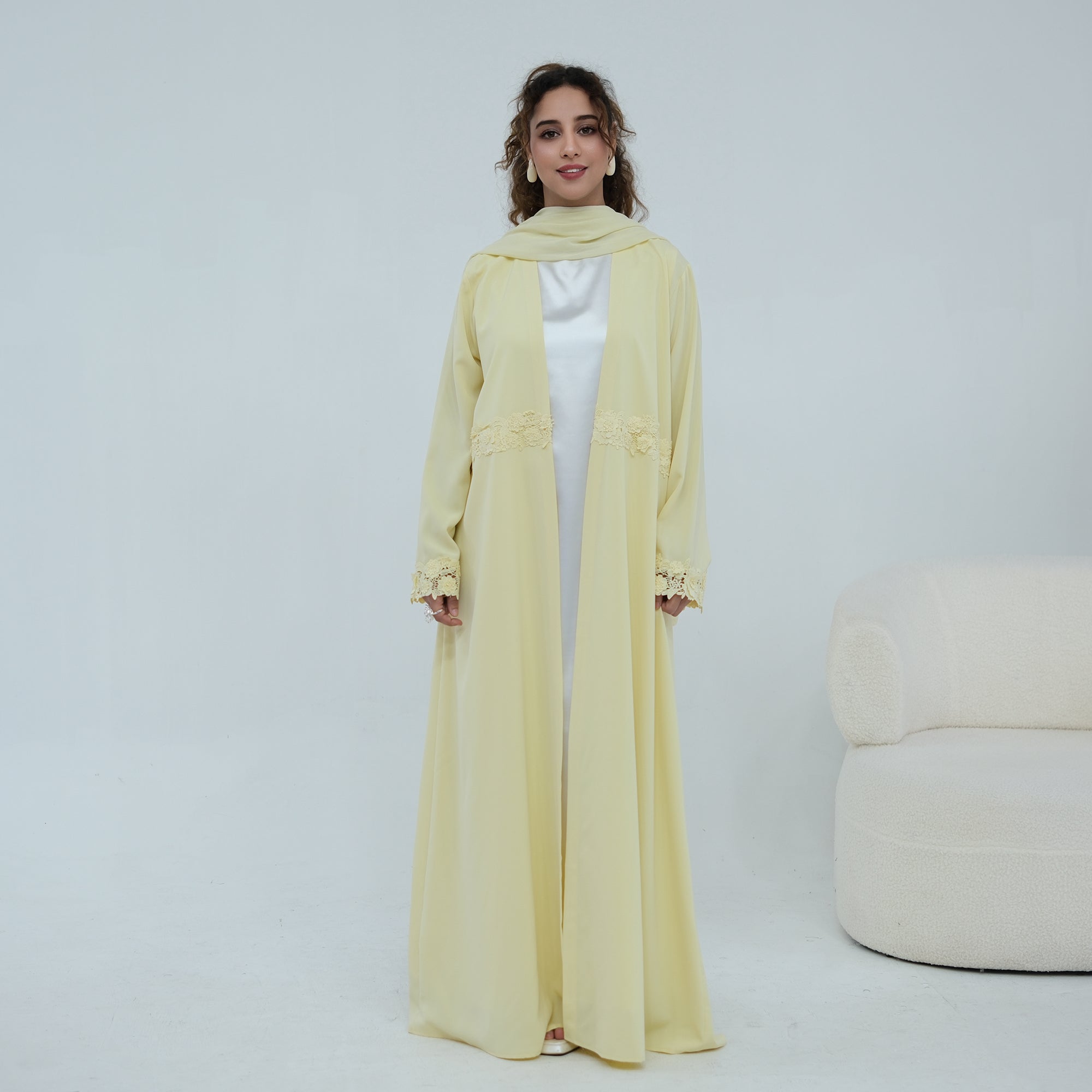 A woman stands in a studio wearing a long, flowing pale yellow abaya with a matching headscarf and a white inner dress visible underneath. A light upholstered chair is partially visible at the right edge.
