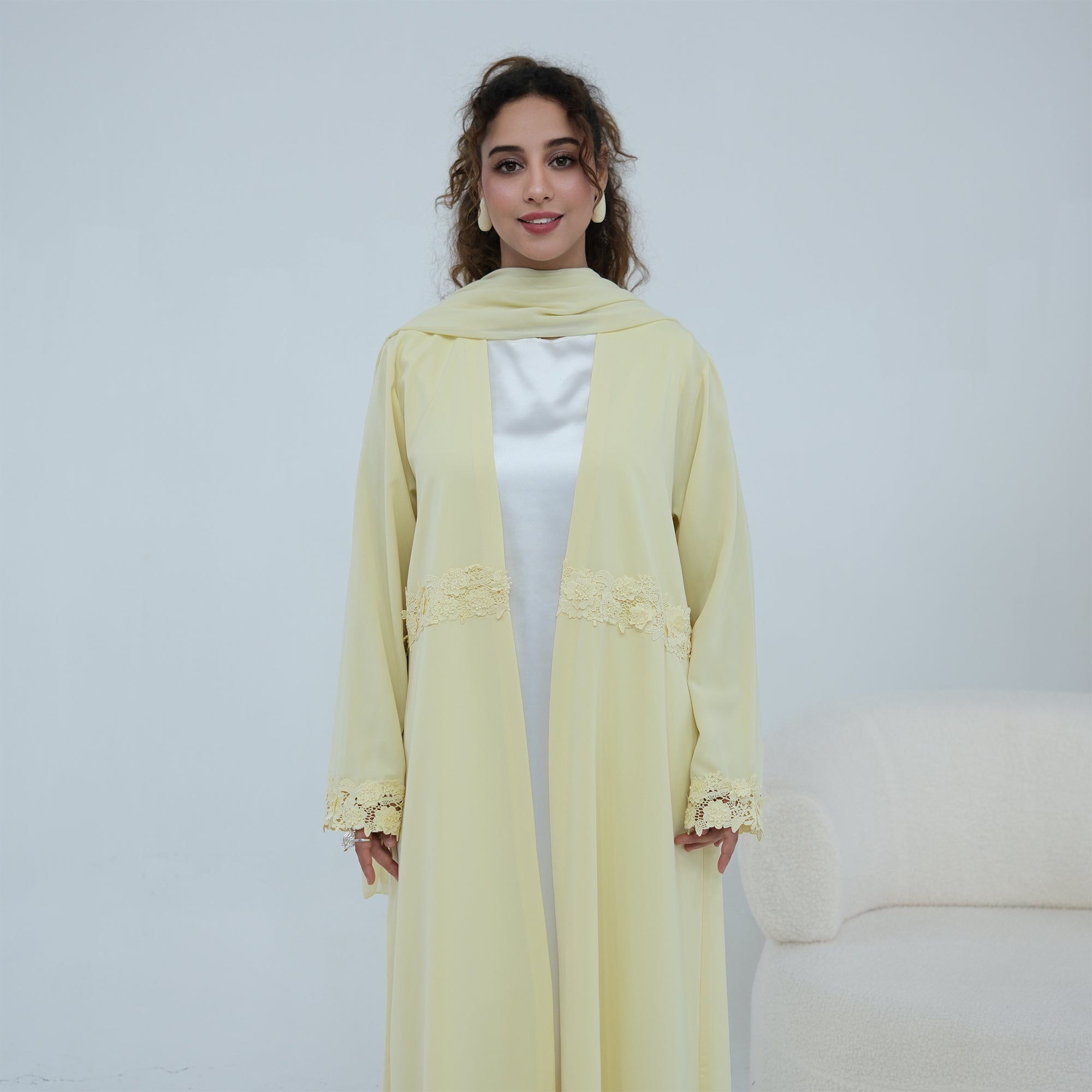 A woman stands wearing a pale yellow long abaya with matching scarf and lace trim details, layered over a white inner dress, in a bright studio setting.