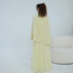 A woman stands with her back to the camera wearing a flowing pale yellow abaya and matching outer layer in a bright, minimalist studio. A cream-colored curved chair is visible on the right side.