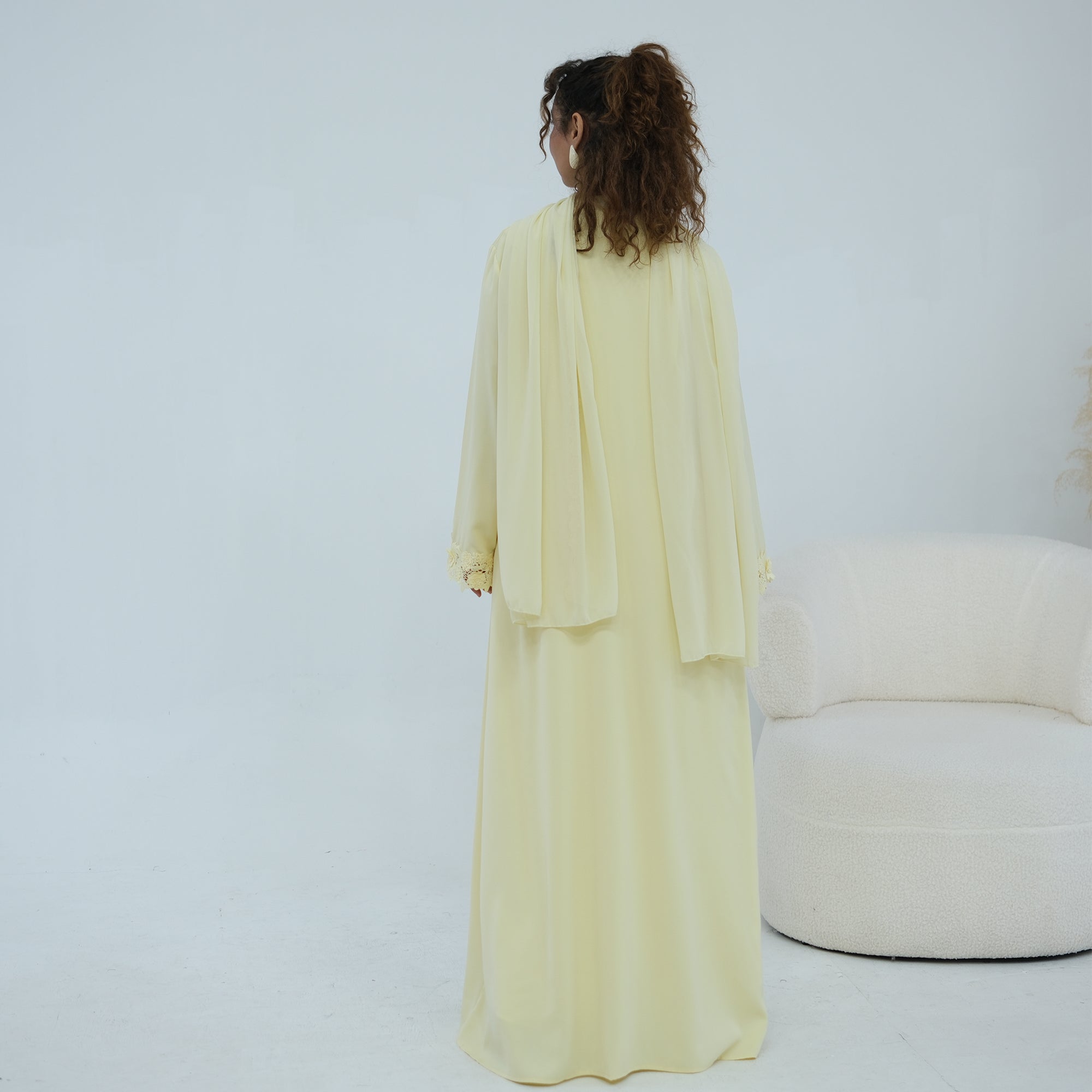 A woman stands with her back to the camera wearing a flowing pale yellow abaya and matching outer layer in a bright, minimalist studio. A cream-colored curved chair is visible on the right side.
