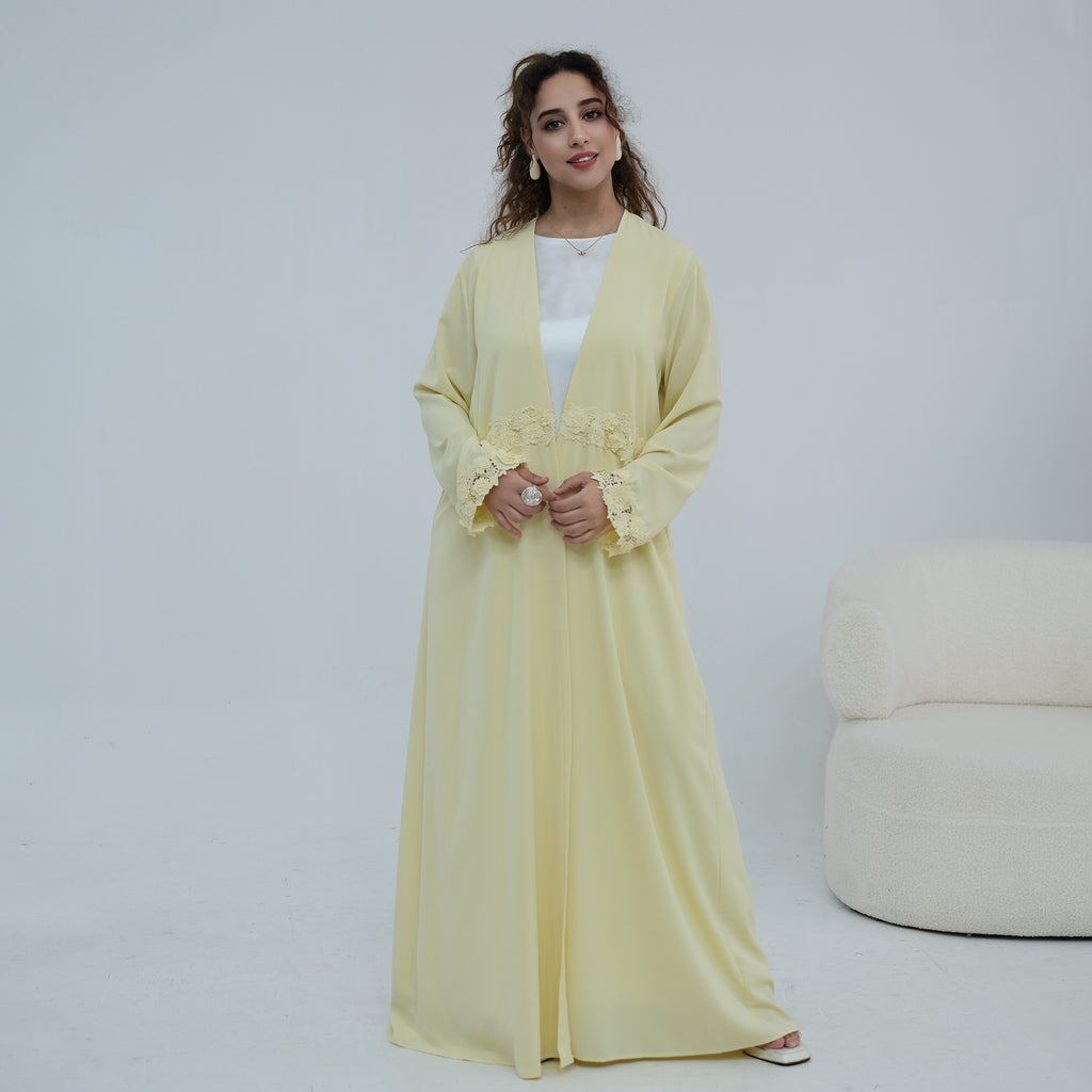 A woman modeling a long, flowing pale yellow abaya with floral lace detailing on the cuffs and waist, worn over a white top in a bright, minimalist studio setting.
